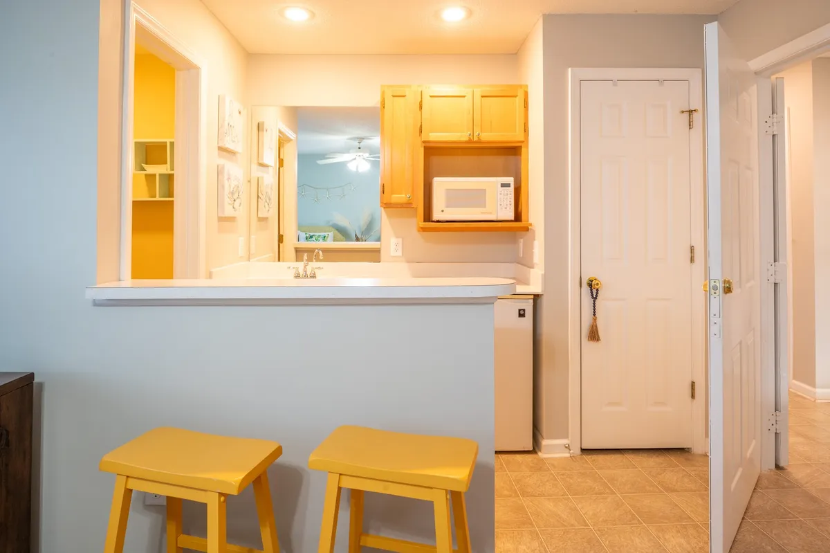 This cozy interior scene features a kitchenette with a kitchen counter, yellow stools for bar seating, a visible microwave, and cabinetry. The area is designed for casual dining or quick snacks, with a welcoming ambience that encourages guests to gather, eat, or chat.
