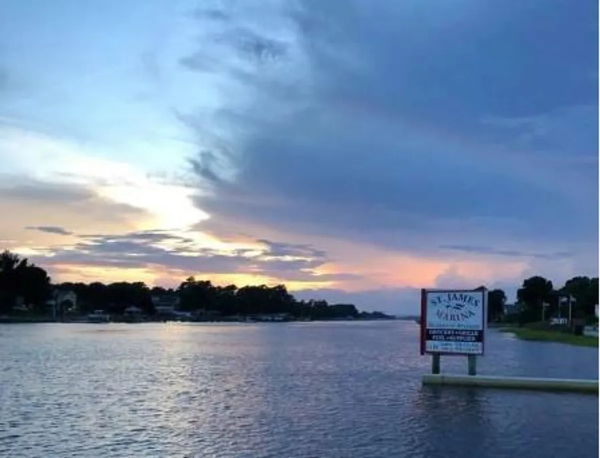 A tranquil river view at sunset, featuring a prominent sign for St. James Marina, calm water, and a colorful sky. The serene setting invites thoughts of peaceful walks and enjoying nature alongside the waterfront.