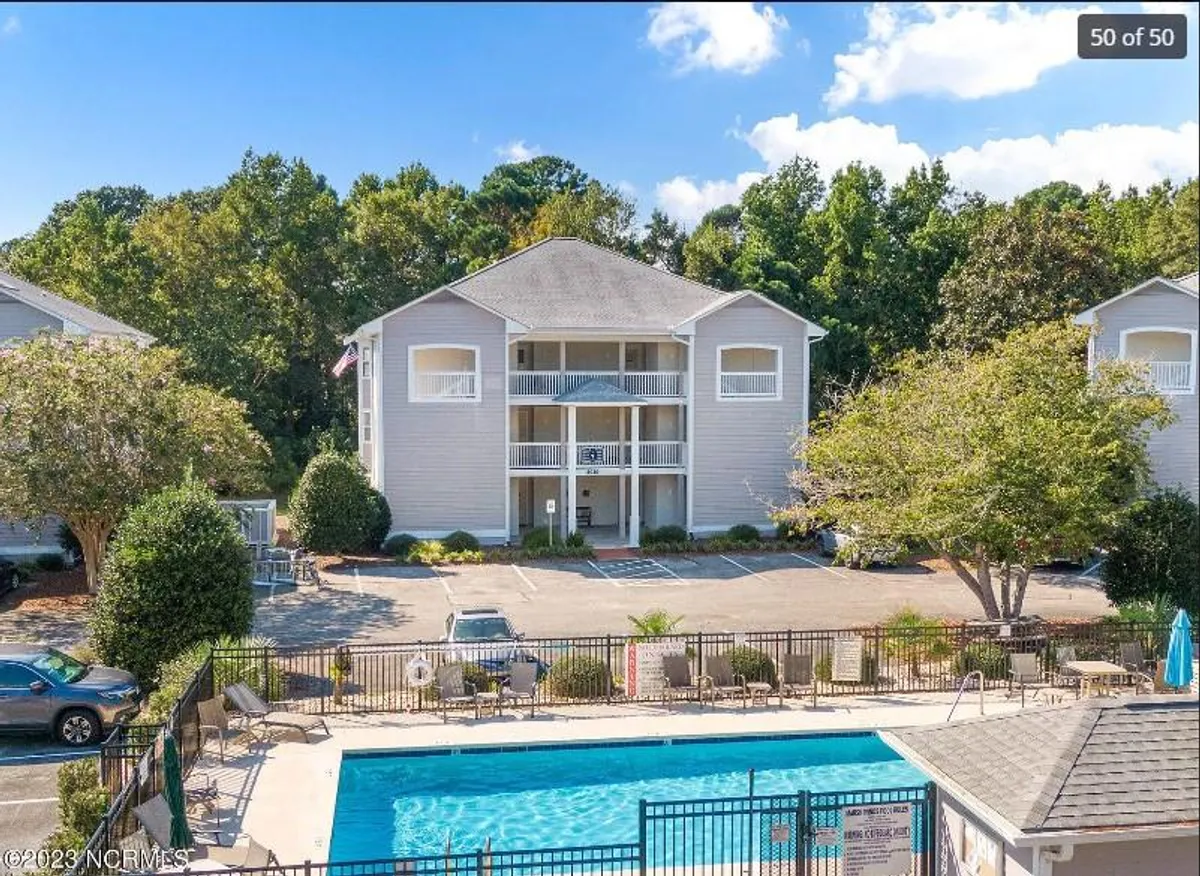 The hero image showcases the inviting exterior of the condo building set within lush green landscaping. The scene centers around a sparkling swimming pool, surrounded by sun loungers and palm trees, creating a relaxing, resort-like atmosphere. The bright sunlight and open skies evoke a sense of tranquility and leisure.