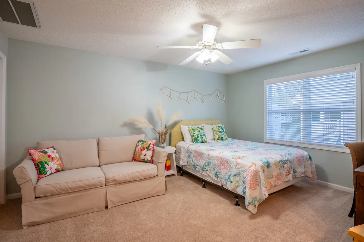 This image features a bed with floral bedding, a full-size sofa with colorful pillows, and a window with blinds. The area looks cozy and cheerful, suggesting both comfort for relaxing and additional sleeping space for guests or family.