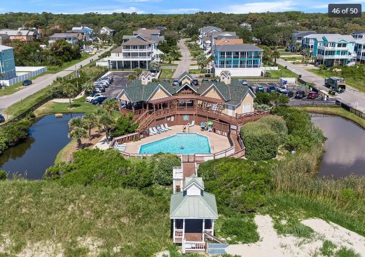 A sparkling swimming pool bordered by a wooden deck and lush greenery, with nearby houses visible in the distance. The setting is communal and inviting, suggesting opportunities for sunbathing, swimming, and socializing in a resort-like atmosphere.