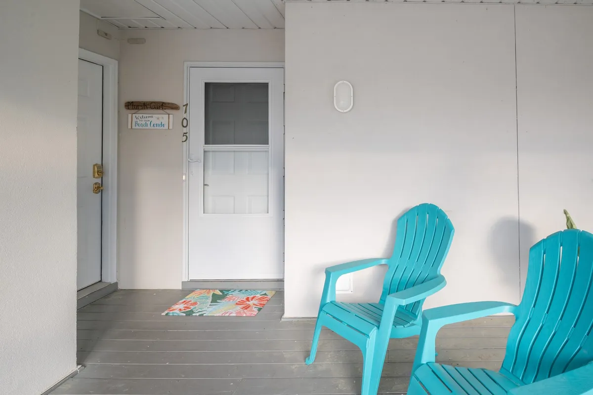 An exterior view of a welcoming porch with turquoise chairs, a visible door marked '705,' a welcome mat, and a sign reading 'Aquarium Beach Condo.' The scene exudes hospitality and a casual, coastal vibe, inviting guests to relax and socialize while enjoying the outdoors.