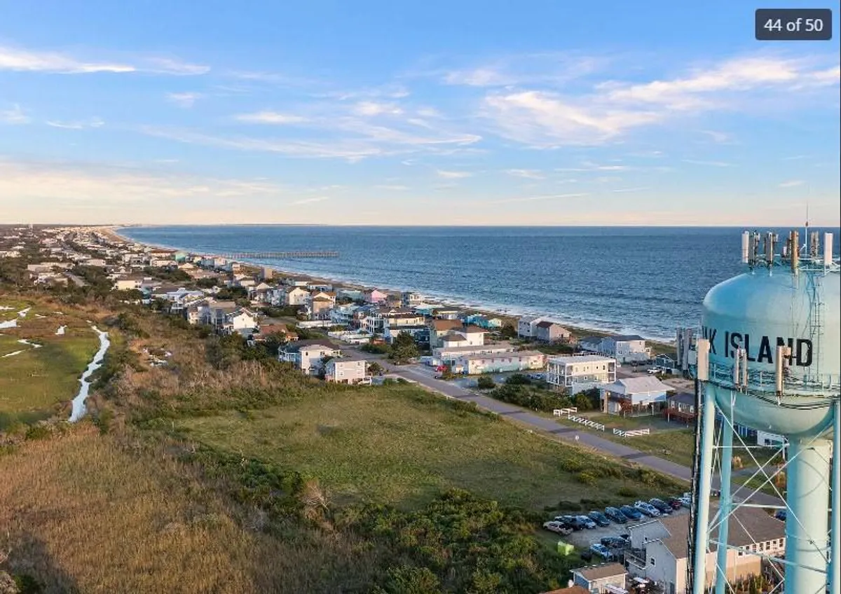 A sweeping, elevated view encompassing the beach, ocean, coastline, and a water tower, with scattered coastal houses in the foreground and lush greenery. The scene captures the expansive shoreline and the tranquil ambiance of the area, hinting at beach walks and the serenity of seaside living.