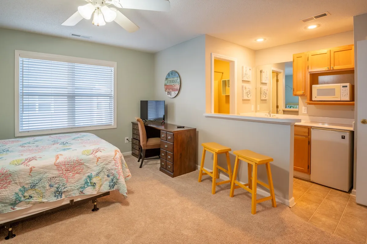 This image showcases a bright and functional studio setup featuring a bed with a colorful quilt, a work desk with a chair, and a compact kitchenette area with bar stools. Natural light fills the space, emphasizing both the comfort of the sleeping area and the practicality of the work and dining zones.