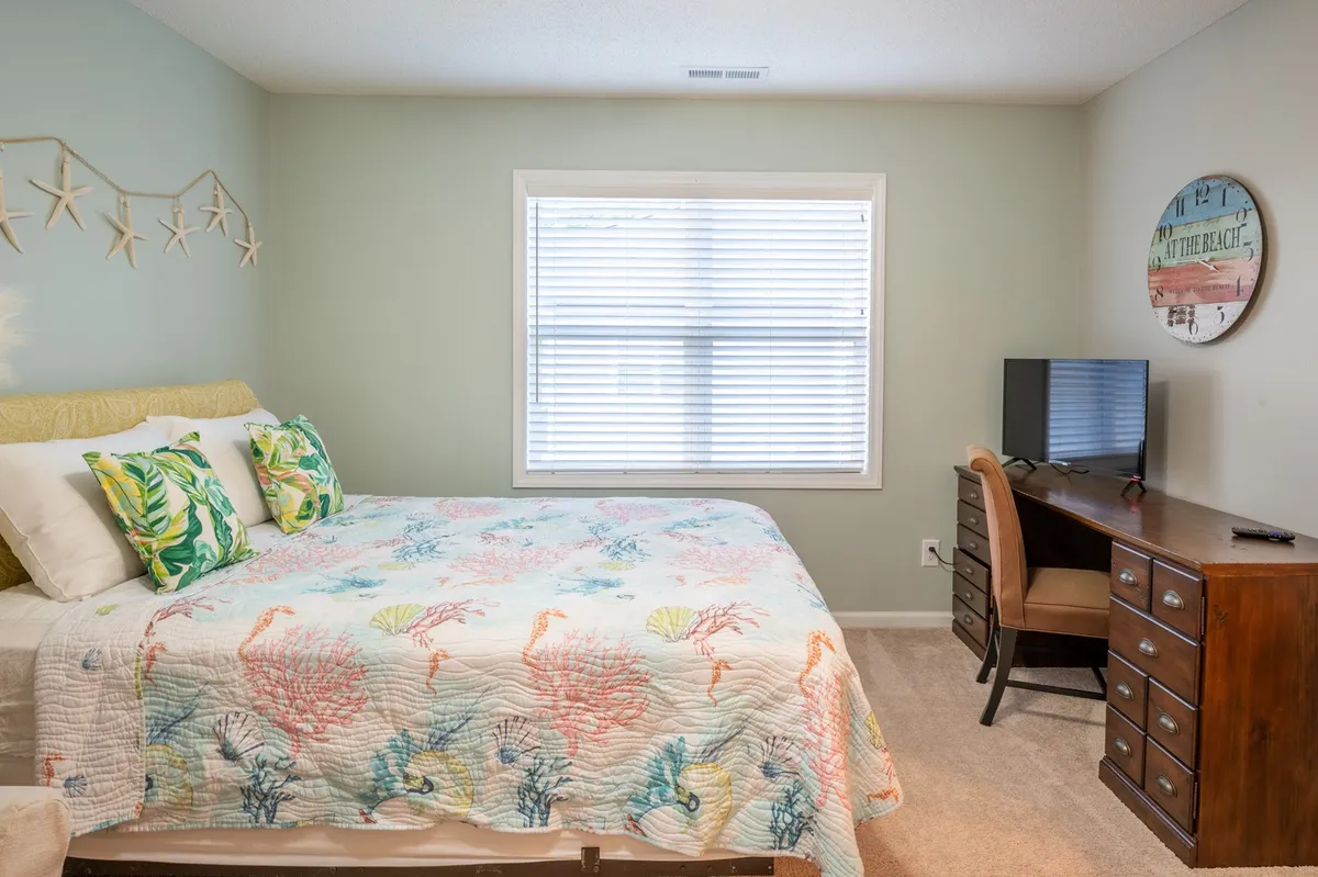 A cozy bedroom setup featuring a bed with a colorful quilt, a desk with a chair, and a window with blinds letting in natural light. The desk offers a welcoming spot for work or reflection, and the overall atmosphere is calm and inviting.